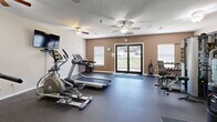 Located at 617 Richardson St in Simpsonville, SC, this fitness center offers a well-equipped exercise space designed to support a variety of workout routines. The room features durable rubber flooring throughout, providing a safe and functional surface for both cardio and strength training activities.

The cardio section is equipped with multiple treadmills and elliptical machines, allowing several users to exercise simultaneously. A multi-station strength training machine, free weights, and a weight bench round out the strength equipment offerings, providing options for full-body workouts. A wall-mounted flat-screen television is positioned for viewing during exercise, and a small utility table provides space for personal items.

Large mirrors line one wall, visually expanding the space and allowing users to monitor their form. Multiple ceiling fans are installed overhead to promote air circulation and comfort during workouts. Natural light fills the room through several windows with blinds and a set of glass double doors that open to the exterior, offering pleasant outdoor views. A water cooler station is conveniently available to keep users hydrated. The warm, neutral-toned walls and practical layout create a clean, focused environment well-suited for daily fitness use.