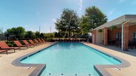 This outdoor pool area is a well-appointed recreational amenity designed for both relaxation and socializing. At its center sits a generously sized swimming pool with crystal-clear blue water, accessible via stainless steel entry rails and steps at multiple points. The pool is finished with a classic tiled border and surrounded by a broad concrete sundeck that provides ample space for lounging.

Lining the perimeter of the pool are rows of rust-colored lounge chairs, offering plenty of seating for sunbathing. Neatly trimmed shrubs and landscaping run along the iron security fence that encloses the entire area, providing both privacy and a polished aesthetic.

Adjacent to the pool is a single-story brick clubhouse building with a covered patio supported by brick columns. The patio is outfitted with round outdoor dining tables and chairs, making it an ideal space for gathering in the shade. Hanging ferns add a decorative touch overhead. A beverage vending machine is conveniently located within the covered area. The open-air setting benefits from abundant natural light and views of mature trees beyond the fence line, contributing to an inviting and comfortable outdoor atmosphere.