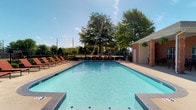 This outdoor pool area is a well-appointed recreational amenity designed for both relaxation and socializing. At its center sits a generously sized swimming pool with crystal-clear blue water, accessible via stainless steel entry rails and steps at multiple points. The pool is finished with a classic tiled border and surrounded by a broad concrete sundeck that provides ample space for lounging.

Lining the perimeter of the pool are rows of rust-colored lounge chairs, offering plenty of seating for sunbathing. Neatly trimmed shrubs and landscaping run along the iron security fence that encloses the entire area, providing both privacy and a polished aesthetic.

Adjacent to the pool is a single-story brick clubhouse building with a covered patio supported by brick columns. The patio is outfitted with round outdoor dining tables and chairs, making it an ideal space for gathering in the shade. Hanging ferns add a decorative touch overhead. A beverage vending machine is conveniently located within the covered area. The open-air setting benefits from abundant natural light and views of mature trees beyond the fence line, contributing to an inviting and comfortable outdoor atmosphere.
