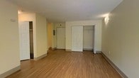 This single-floor space is designed with simplicity and practicality in mind. The main living area is finished with warm hardwood-style laminate flooring that extends throughout, providing a cohesive and easy-to-maintain surface underfoot. Neutral cream-toned walls keep the interior feeling bright and open, offering a versatile backdrop for a variety of furnishing styles.

A prominent double-door closet is a standout feature of the main room, providing generous built-in storage with an open interior visible through the partially ajar doors. The closet's white paneled doors add a clean, classic detail to the space. A hallway visible in the background leads further into the unit, suggesting additional rooms or utility areas beyond the main living space. Wall-mounted lighting fixtures provide supplemental illumination throughout.

The unit's electrical panel is conveniently accessible within the living area, and baseboards line the perimeter, contributing to the well-maintained appearance of the space. Overall, the unit presents a functional, move-in-ready environment with a straightforward, no-frills aesthetic suited to a range of occupants.