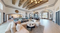 This expansive single-floor space is defined by its striking architectural character and sophisticated interior design. Soaring ceilings with exposed rustic wood beams create a grand sense of volume, while bold crown molding trim lines the upper walls, adding refined structure to the open layout. The space flows seamlessly between distinct living and kitchen areas, connected by a series of elegant arched doorways framed in dark charcoal — a signature design detail that repeats throughout.

The living area features wide-plank light wood flooring, large-scale sectional seating, and a curated mix of contemporary artwork, geometric wall shelving in navy blue, and a stone accent fireplace wall. Designer pendant lighting and globe chandeliers provide warm, layered illumination throughout. Decorative elements, including abstract wall sculptures, oversized clocks, and metallic wall accents, add visual interest at every turn.

The kitchen is appointed with granite countertops, gray-blue cabinetry, stainless steel appliances, and dramatic gold globe pendant lights that anchor the central island. Floor-to-ceiling windows along the perimeter flood the space with abundant natural light and offer views of the outdoor surroundings, including what appears to be a resort-style pool area. The overall ambiance is modern, warm, and thoughtfully curated.