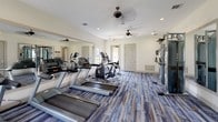 Located at 20900 FM-1093 W in Katy, Texas, this single-floor fitness center is a well-appointed exercise facility designed to support a full range of workout routines. The space features a bright, open layout with crisp white walls, recessed lighting, and ceiling fans that maintain a comfortable atmosphere throughout.

The flooring throughout the fitness center is a distinctive blue-toned wood-look material that adds a modern, stylish character to the space. Large, multi-pane windows line the walls, allowing natural light to pour in and offering views of the surrounding landscaped grounds, creating an energizing environment for exercise.

The cardio area is equipped with multiple treadmills, elliptical machines, and a recumbent bike, providing ample options for aerobic training. Full-length wall mirrors are strategically placed to maximize the sense of space and assist with form during workouts. A multi-station cable machine supports strength training needs, while a dedicated free weights area features a well-stocked dumbbell rack.

An additional corner of the fitness center is dedicated to group fitness accessories, including stability balls, medicine balls, and a Smart Fitness rack, complemented by a wall-mounted flat-screen television. Multiple access doors throughout the space ensure easy entry and exit. Overall, this fitness center offers a clean, functional, and inviting environment for residents to pursue their health and wellness goals.