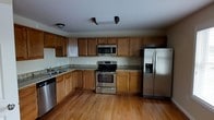 This single-floor space showcases a functional and well-appointed kitchen as its centerpiece. The kitchen is designed with an L-shaped layout, offering ample counter space finished with granite-style countertops that provide both durability and visual appeal. Warm-toned wood cabinetry lines the walls, extending from floor to ceiling to maximize storage capacity, with upper and lower cabinets providing generous organizational options throughout the space.

The kitchen is equipped with a full suite of stainless steel appliances, including a freestanding electric range with oven, an over-the-range microwave, a side-by-side refrigerator with an integrated ice and water dispenser, and a dishwasher. A sink with a single-basin setup is positioned along the counter, complementing the practical workflow of the kitchen.

The flooring throughout the space consists of warm-toned hardwood, adding a classic and inviting character to the interior. Overhead lighting illuminates the kitchen efficiently. The open feel of the kitchen, combined with the natural wood tones of the cabinetry and flooring, creates a cohesive and welcoming ambiance suited for both everyday use and entertaining.