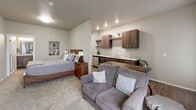 This efficiently designed single-floor unit offers a thoughtful open-concept layout that seamlessly integrates the living, sleeping, and kitchen areas into one cohesive space. The main living area features warm, neutral-toned carpet that transitions to rich hardwood-style flooring in the kitchen corridor, creating a natural visual distinction between zones.

The kitchenette is outfitted with dark espresso cabinetry, granite-look countertops, a stainless steel sink, a microwave, and a dishwasher. Open upper shelving adds a modern touch while keeping the space feeling airy. A small dining bar with seating is positioned nearby, making it functional for everyday use.

The sleeping area is generously sized relative to the overall footprint, accommodating a full bed with a solid wood headboard, a nightstand, and a cozy seating area. A window allows natural light to brighten the room, while recessed lighting provides consistent illumination throughout.

The bathroom features a dark wood vanity with a granite-style countertop, a framed mirror, tile flooring, and a shower with a curtain enclosure. The dedicated laundry room is a standout practical amenity, housing a side-by-side washer and dryer unit with overhead shelving for additional storage. Neutral wall tones and consistent finishes throughout lend the space a clean, modern, and welcoming ambiance.