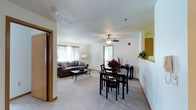 This single-floor space features an open-concept layout that seamlessly connects the living and dining areas, creating a cohesive and functional environment. The living area is carpeted and comfortably sized, accommodating a sofa and coffee table beneath a ceiling fan with a light fixture, which serves as a central focal point overhead. Natural light filters in through a curtained window, brightening the neutral-toned walls and contributing to a welcoming ambiance.

Adjacent to the living area, the dining space is defined by a dark wood table and matching chairs, providing a dedicated area for meals. A decorative floral arrangement on the table adds a touch of warmth. The transition between the two zones flows naturally without visual barriers, enhancing the sense of openness throughout.

A passageway visible from the dining area leads to additional rooms, while a built-in shelf accent adds a subtle architectural detail along one wall. The textured ceiling and light, neutral color palette throughout lend the space a clean, understated character. Overall, the layout prioritizes ease of movement and practical use of the available space.