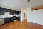This single-floor residence showcases a thoughtfully designed interior with a cohesive blend of contemporary finishes throughout. Rich hardwood-style flooring flows continuously across the open-concept living and kitchen areas, creating a warm, unified aesthetic.

The kitchen is a standout feature, outfitted with deep navy-toned shaker cabinetry accented by gold hardware and complemented by striking granite countertops. A herringbone-patterned tile backsplash adds visual texture, while a full suite of stainless steel appliances—including a refrigerator, range, microwave, and dishwasher—completes the space.

The open living area is generously proportioned, featuring recessed lighting and a ceiling fan. A uniquely positioned clerestory cutout above connects visual interest between spaces.

The hallway leads to additional rooms via a combination of panel doors and a sleek sliding barn door fitted with black metal hardware—a modern farmhouse detail that adds character. The bedroom benefits from multiple large windows that invite abundant natural light and outdoor views.

The bathroom is finished with neutral large-format wall tiles surrounding a soaking tub, matte black fixtures, and dark vanity cabinetry with granite countertops, maintaining the consistent design language found throughout the residence.