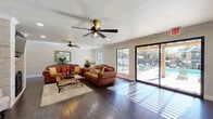 This well-appointed single-story home showcases a thoughtful, open layout united by rich dark hardwood flooring throughout. The expansive living room serves as the heart of the home, anchored by a striking whitewashed brick fireplace and complemented by dual ceiling fans, crown molding, and a textured ceiling. Generous natural light pours in through large windows and sliding glass doors that offer direct views and access to a covered outdoor patio and sparkling pool area beyond, seamlessly blending indoor and outdoor living.

Adjacent to the living room, a dedicated sitting area provides a quieter retreat, featuring an exposed brick accent wall, elegant upholstered chairs, and warm neutral tones. The dining area is bright and inviting, with a substantial wood dining table and large windows framing lush outdoor scenery, perfect for gatherings.

The kitchen is spacious and functional, designed in a classic U-shape with abundant white cabinetry, contrasting matte black hardware, a warm earth-toned subway tile backsplash, light countertops, recessed lighting, and stainless steel appliances. Together, the spaces create a cohesive, comfortable home that balances casual warmth with practical design, ideal for everyday living and entertaining alike.