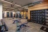 This fitness center in San Antonio, TX offers a spacious, single-floor layout designed to support a wide variety of workout styles. The interior features an industrial-modern aesthetic, with exposed ductwork running along the ceiling, sleek pendant lighting, and ceiling fans that complement the contemporary design. Patterned carpet flooring with teal accent strips guides movement throughout the facility, creating distinct workout zones. Large windows along one wall flood the cardio area with natural light and provide views of the surrounding outdoor space, where treadmills, stationary bikes, and rowing machines are positioned to take full advantage of the outlook. The strength training area is outfitted with a comprehensive dumbbell rack, adjustable benches, a cable multi-station machine, and plate-loaded equipment. A functional training zone houses battle ropes, kettlebells, medicine balls, a rig system, and a BOSU balance trainer, all reflected in large floor-to-ceiling mirrors that enhance the sense of space. Motivational wall graphics and branded signage contribute to an energetic atmosphere throughout. The facility is well-lit with linear LED fixtures and includes wall-mounted TVs, lockers near the entry, and clearly marked exits, ensuring both comfort and safety for all users.