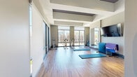 Located at 2007 Knoll Top Ln in Franklin, TN, this well-appointed fitness studio offers a bright and functional space designed to support a variety of workout routines. The room features warm hardwood-style flooring throughout, complemented by a distinctive tray ceiling that adds architectural depth and a sense of height to the interior.

Large, floor-to-ceiling windows line one wall of the studio, flooding the space with abundant natural light and providing open views of the surrounding Franklin streetscape. Roller shades offer the option to control light levels as needed. The expansive glazing also connects the studio visually to an adjacent multi-story atrium area, reinforcing a sense of openness.

The studio is equipped with stationary cycling bikes, exercise mats, a mini trampoline, a dumbbell and equipment rack, and additional cardio and strength apparatus. A large wall-mounted mirror enhances the sense of spaciousness and supports form-checking during workouts, while a flat-screen television mounted on the opposite wall adds an entertainment option. The neutral taupe and white color palette keeps the environment clean and focused. Access to what appears to be an additional fitness or weight room is visible through a glass-paneled door, suggesting further amenities nearby. Overall, the studio delivers a practical and polished environment for diverse fitness activities.