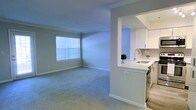 This single-floor residence offers a clean, open-concept layout designed for comfortable everyday living. The spacious living area is finished with soft wall-to-wall carpeting that adds warmth underfoot, while neutral-toned walls and crown molding lend a polished, cohesive aesthetic throughout. A large window fitted with horizontal blinds floods the living space with natural light, creating a bright and airy atmosphere.

A defining architectural feature is the pass-through opening between the living area and the kitchen, which promotes an open, connected feel while maintaining a subtle sense of separation between the two spaces. The kitchen is outfitted with crisp white upper cabinetry, a light-toned countertop, and a stainless steel sink. A tile backsplash adds a touch of texture and detail to the kitchen's clean, modern finish. The flooring transitions from carpet in the living area to hard surface in the kitchen, providing both practicality and visual contrast.

Crown molding runs consistently throughout the visible spaces, reinforcing a well-finished interior. The overall ambiance is light, neutral, and move-in ready, with a design that balances simplicity with thoughtful detail — well-suited for those seeking a tidy, functional, and welcoming living environment.