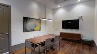 This single-floor space showcases a thoughtfully designed interior centered around a rich, warm-toned wooden conference or dining table surrounded by contemporary gray upholstered chairs with polished chrome legs. The room's most striking architectural feature is its soaring ceiling height, which contributes to an open, airy atmosphere. A linear pendant light fixture hangs above the table, providing focused illumination and serving as a design focal point.

The walls are finished in a clean, neutral white that enhances the sense of brightness and space. Recessed lighting supplements the pendant fixture, ensuring even light distribution throughout the room. A large flat-screen television is wall-mounted on one side, supported by a sleek mid-century-style wooden media console, adding both functionality and visual warmth. On the opposite wall, a large-scale abstract painting in muted greens and yellows introduces an artistic element and a subtle pop of color.

The flooring consists of rich hardwood planks in a deep, warm tone, grounding the space and complementing the wooden furnishings. Overall, the space exudes a refined, modern aesthetic suitable for professional meetings or elegant gatherings.