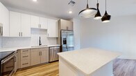 This single-floor unit offers a thoughtfully designed layout across a compact yet efficient footprint. The kitchen serves as a standout feature, showcasing two-tone cabinetry with white upper cabinets and light wood-grain lower cabinets, complemented by a classic white subway tile backsplash and light quartz countertops. A suite of stainless steel appliances—including a French door refrigerator, range, built-in microwave, and dishwasher—provides full functionality, while a central island with a matching quartz surface adds additional workspace and casual dining potential. Matte black pendant lights and cabinet hardware add a modern contrast throughout. Light hardwood-style flooring runs through the main living and kitchen areas, contributing to a bright, airy atmosphere.

The bedroom is carpeted for comfort and includes a ceiling fan, a natural light-providing window with blinds, and a walk-in closet, offering practical storage. The bathroom features large-format tile flooring, a wide vanity with quartz countertop and black hardware, an illuminated frameless mirror, and a tub-shower combination with tiled surround. The overall design is clean, contemporary, and efficiently organized.