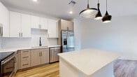 This single-floor unit offers a thoughtfully designed layout across a compact yet efficient footprint. The kitchen serves as a standout feature, showcasing two-tone cabinetry with white upper cabinets and light wood-grain lower cabinets, complemented by a classic white subway tile backsplash and light quartz countertops. A suite of stainless steel appliances—including a French door refrigerator, range, built-in microwave, and dishwasher—provides full functionality, while a central island with a matching quartz surface adds additional workspace and casual dining potential. Matte black pendant lights and cabinet hardware add a modern contrast throughout. Light hardwood-style flooring runs through the main living and kitchen areas, contributing to a bright, airy atmosphere.

The bedroom is carpeted for comfort and includes a ceiling fan, a natural light-providing window with blinds, and a walk-in closet, offering practical storage. The bathroom features large-format tile flooring, a wide vanity with quartz countertop and black hardware, an illuminated frameless mirror, and a tub-shower combination with tiled surround. The overall design is clean, contemporary, and efficiently organized.
