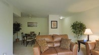 This single-floor residence offers a thoughtfully arranged living space defined by a warm, neutral color palette and an open-concept layout that seamlessly connects the main living and dining areas. The living area is anchored by a plush brown leather loveseat accented with decorative throw pillows, creating an inviting space for relaxation. Soft, light-toned carpeting runs throughout, contributing to the room's cozy and understated aesthetic. A ceiling fan overhead provides both functionality and a subtle design element to the space. Adjacent to the living area, the dining space accommodates a dark wood dining table with a matching set of chairs — ideal for everyday meals or casual gatherings. Framed wall art adds character and a personal touch to the space, while lush indoor greenery introduces a refreshing natural element. The clean, white walls and consistent lighting throughout give the space a bright and airy feel. The open flow between living and dining zones enhances the sense of spaciousness, making this single-floor layout both practical and comfortable for everyday living.