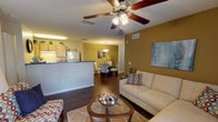 This single-floor residence offers a thoughtfully designed layout that balances comfort and functionality. The open-concept living area serves as the heart of the home, featuring rich hardwood-style flooring, warm earth-toned walls, and a ceiling fan with integrated lighting. A large sliding glass door floods the space with natural light and provides access to the outdoors. The living area flows seamlessly into a dedicated dining space, illuminated by a pendant light fixture and anchored by neutral-toned walls that create a calm, welcoming atmosphere.

The galley-style kitchen is well-equipped, featuring light wood cabinetry, dark countertops, a double basin sink, and white appliances including an electric range and refrigerator. A built-in pantry closet provides generous storage space.

The residence includes two carpeted bedrooms, each featuring ceiling fans, ample closet space, and softly neutral wall tones that contribute to a restful environment. Natural light filters through the windows, enhancing the tranquil feel of each room.

The full bathroom is finished with wood-toned cabinetry, a single vanity with a wall-mounted mirror, overhead lighting, and a shower-tub combination. Hardwood-style flooring extends into the bathroom, maintaining a cohesive aesthetic throughout the home.