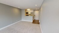 This single-floor unit features a practical and well-organized layout designed to maximize usable space. The living area is generously proportioned, finished with soft neutral carpet and painted in a calming light gray palette throughout. Recessed lighting and a textured ceiling provide a clean, contemporary look, while a large sliding glass door floods the room with natural light and opens to a private outdoor area with views of surrounding greenery. Vertical blinds offer flexible light control.

The kitchen is positioned off the main living space behind a peninsula-style breakfast bar, creating an open connection between the two areas. White cabinetry lines both walls of the galley-style kitchen, complemented by granite-patterned countertops, a stainless steel double sink, a gas range, and a stainless steel over-the-range microwave. Wood-look vinyl flooring runs throughout the kitchen and hallway areas, providing a durable contrast to the carpeted living space.

The bathroom includes a white vanity with cabinet storage, a wall-mounted mirror with overhead lighting, and a shower/tub combination. An adjacent closet offers additional storage. The overall design presents a cohesive, move-in-ready space with consistent finishes and a neutral color scheme throughout.