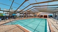 This outdoor amenity space centers around a large, rectangular swimming pool encased within a distinctive barrel-vaulted screen enclosure supported by a structural metal and wood frame. The translucent screening allows abundant natural light to flood the deck while providing protection from the elements, creating a bright and airy atmosphere ideal for year-round enjoyment. The pool features clear turquoise water, underwater tiling in a grid pattern, and entry steps with metal handrails for easy access. Adjacent to the main pool is a raised spa or hot tub, accessible via brick paver steps. The surrounding deck is finished with attractive brick pavers and is generously sized, accommodating multiple blue lounge chairs and seating arrangements positioned for sunbathing and relaxation. A shaded seating area with a blue umbrella and outdoor furniture provides a comfortable retreat. Lush greenery and mature trees are visible beyond the enclosure, and views of the surrounding multi-story residential buildings frame the space. The overall ambiance is resort-like, combining open-air brightness with the comfort of a sheltered environment, making it a standout communal feature of the residential community.