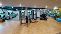 Located at 2200 W Meeker St in Kent, Washington, this single-floor fitness center offers a comprehensive workout environment designed to accommodate a wide range of exercise routines. The space is organized into distinct functional zones, each tailored to a specific type of training.

The cardio area features a row of treadmills and other cardio machines positioned along large windows that frame views of the surrounding neighborhood, flooding the zone with natural light and creating an open, energizing atmosphere. The dark-painted exposed ceiling with industrial ductwork and recessed lighting adds a modern, urban aesthetic to this section.

The strength training zone is equipped with multiple cable and weight machines, adjustable benches, and a well-stocked dumbbell rack spanning a range of weights. Full-length mirrors line the walls, visually expanding the space and supporting proper form during workouts.

A dedicated free weights and functional training area includes barbells, weight plates, medicine balls, stability balls, and a squat rack, offering ample space for dynamic movement. Throughout the facility, light wood-toned flooring provides a clean, cohesive look, while bold, abstract graphic murals in teal, black, and white serve as striking visual focal points, reinforcing the space's contemporary and energetic character.