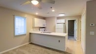 This single-floor residence offers a clean, functional layout centered around an open-concept kitchen and living area. The kitchen is well-appointed with white cabinetry that extends to the upper walls, providing generous storage. A built-in microwave and freestanding range sit neatly along the kitchen wall, while a full-size refrigerator anchors the far corner. The kitchen features a wraparound peninsula with a light-colored countertop, offering additional workspace and a natural boundary between the kitchen and the adjoining living area. The living space is finished with neutral carpet and light-toned walls that lend a calm, airy atmosphere throughout. Recessed lighting and a ceiling fan with an integrated light fixture provide both ambient illumination and practical air circulation. Natural light filters in through a window fitted with horizontal blinds, brightening the space during daylight hours. A doorway leads from the main living area into an additional room, suggesting a flexible floor plan with space for multiple uses. The overall design is straightforward and move-in ready, emphasizing practicality and a cohesive neutral palette throughout.