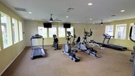 This single-floor fitness center offers a spacious, open layout designed to accommodate a comprehensive workout experience. The room features warm-toned walls paired with neutral carpeted flooring, creating a welcoming and energizing environment for exercise. Recessed lighting throughout the ceiling ensures the space is well-lit, while ceiling fans provide comfortable airflow during workouts. Large windows along one wall allow generous natural light to fill the room, offering views of the surrounding outdoor area and keeping the atmosphere bright and open. Wall-mounted flat-screen televisions are positioned at eye level, allowing users to stay entertained during cardio sessions. The fitness floor is equipped with a diverse selection of exercise machines, including treadmills, elliptical trainers, upright and recumbent stationary bikes, and a multi-function strength training station, as well as additional resistance equipment. The machines are thoughtfully arranged to allow for ease of movement and accessibility throughout the space. The overall ambiance is clean, functional, and purposefully designed to support a variety of fitness routines in a comfortable, community-oriented setting.