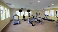 This single-floor fitness center offers a spacious, open layout designed to accommodate a comprehensive workout experience. The room features warm-toned walls paired with neutral carpeted flooring, creating a welcoming and energizing environment for exercise. Recessed lighting throughout the ceiling ensures the space is well-lit, while ceiling fans provide comfortable airflow during workouts. Large windows along one wall allow generous natural light to fill the room, offering views of the surrounding outdoor area and keeping the atmosphere bright and open. Wall-mounted flat-screen televisions are positioned at eye level, allowing users to stay entertained during cardio sessions. The fitness floor is equipped with a diverse selection of exercise machines, including treadmills, elliptical trainers, upright and recumbent stationary bikes, and a multi-function strength training station, as well as additional resistance equipment. The machines are thoughtfully arranged to allow for ease of movement and accessibility throughout the space. The overall ambiance is clean, functional, and purposefully designed to support a variety of fitness routines in a comfortable, community-oriented setting.