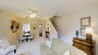 Located at 2545 Croquet Dr in Wilmington, North Carolina, this two-story home offers a comfortable and functional layout spread across two well-connected floors. The main level centers around an open living area finished with soft, neutral-toned carpeting that runs throughout, creating a warm and cohesive atmosphere. A ceiling fan with an integrated light fixture provides both comfort and illumination to the living space, while a coordinating pendant chandelier highlights the adjacent dining area, which is furnished with a round table and upholstered seating. The dining space is positioned near a window, allowing natural light to filter into the room. A staircase with clean white balusters and a wood handrail leads to the upper floor, serving as a functional focal point in the main living area. A doorway visible from the living room suggests additional rooms beyond, with a secondary entry or utility area adding to the practical flow of the main level. Warm wall tones in a soft cream finish lend the interior a bright, inviting character throughout. The overall layout and design reflect a home that balances everyday livability with comfortable, classic detailing suited to the Wilmington, NC lifestyle.