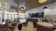 This single-floor community clubhouse is a thoughtfully designed social hub that blends modern coastal aesthetics with comfortable, functional living spaces. The open-plan layout is anchored by wide-plank wood-look flooring that flows throughout, lending warmth and cohesion to the entire space.

The main lounge area showcases a striking wood-paneled ceiling with recessed lighting and bold black dome pendant fixtures. Comfortable seating — including a neutral sofa, eclectic rattan and bamboo chairs, and organic driftwood accent tables — is arranged around a wall-mounted television, creating an inviting gathering spot. A full wall of French doors floods the room with natural light and provides direct access to an outdoor area.

Adjacent to the lounge, a dedicated games zone features a pool table and a regulation shuffleboard table, offering entertainment options alongside a round table for casual seating. A large coastal-themed artwork adds visual character to this area.

The coffee bar serves as a central social point, equipped with a Starbucks coffee machine and sleek gray cabinetry set against a dramatic dark elongated tile backsplash. A generous quartz-topped island with bar-height seating provides space for casual conversation, while pendant globe lighting adds an upscale touch.

Partially enclosed lounge nooks with upholstered booth seating and chevron accent walls create more intimate settings within the broader open space, making this clubhouse equally suited for lively gatherings or quiet relaxation.