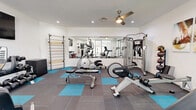 Located at 3501 E Camelback Rd in Phoenix, Arizona, this fitness center offers a thoughtfully organized single-room workout space designed to maximize functionality. The room features crisp white walls, recessed lighting, and a ceiling fan that keeps the environment comfortable during workouts. A large wall-to-wall mirror along one side visually expands the space and allows for form-checking during exercise.

The flooring consists of a modern combination of gray and teal carpet tiles, adding both a vibrant aesthetic touch and a practical surface for varied activities. Equipment is well-rounded and includes a treadmill, elliptical, rowing machine, recumbent bike, and a multi-station cable machine for strength training. A wooden wall-mounted stall bar provides options for stretching and bodyweight exercises, while a dumbbell rack, medicine ball storage, and stability balls round out the free weight and functional fitness offerings.

A wall-mounted television provides entertainment during cardio sessions, and motivational fitness artwork decorates the walls. Glass sliding doors on one side of the room allow natural light to filter in. Posted fitness rules and guidelines reflect an organized, community-conscious environment. The space is efficiently arranged to accommodate a variety of training styles within a compact footprint.