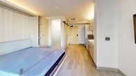 Located on the sixth floor of a modern building in San Francisco's Tenderloin neighborhood, this efficiently designed one-bedroom, one-bathroom unit maximizes every inch of its compact footprint. The open layout integrates the living and sleeping area into a single, well-organized space anchored by a built-in Murphy bed system with integrated shelving and cabinetry in light wood tones, allowing the room to serve multiple functions throughout the day. Light hardwood flooring runs throughout, contributing to a bright, cohesive aesthetic.

The kitchenette runs along one wall and features marble-look quartz countertops, light wood lower cabinetry, gray upper cabinets extending to the ceiling, a stainless steel sink with a matte black faucet, a dishwasher, microwave, and compact refrigerator. A floor-to-ceiling sliding glass door floods the main space with natural light and provides views of the surrounding urban streetscape.

The bathroom is finished in clean white tones with large-format tile flooring and walls. It includes a wall-mounted sink with matte black fixtures, a mirrored medicine cabinet, a wall-mounted toilet, and an open roll-in shower with grab bars, offering accessibility-friendly design. A sliding barn-style door with a matte black pull completes the modern interior detailing throughout the unit.