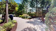 Located at 619 Adams St in Hoboken, New Jersey, this thoughtfully designed outdoor courtyard offers a welcoming retreat nestled between multi-story brick residential buildings. The space is anchored by a herringbone-patterned brick paver pathway that winds through meticulously maintained garden beds, filled with a rich variety of plantings including hostas, ornamental shrubs, Japanese maples, and mature evergreen trees. Overhead, strands of festoon string lights are suspended between posts, adding a charming ambiance to the walkway. The entry is framed by a sleek, modern metal gate that provides a secure yet elegant transition from the street. A dedicated children's playground area is enclosed by chain-link fencing and features a full play structure with slides and climbing equipment, as well as youth sports accessories, all set on a cushioned rubber surface for safety. The surrounding greenery — including tall arborvitae, blue spruce, and deciduous trees in seasonal color — creates a sense of privacy and tranquility within an urban setting. Seating areas with umbrellas are integrated into the landscape, offering comfortable spaces for outdoor relaxation. The overall design balances vibrant horticulture with functional amenities, making it an inviting community outdoor space.