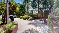 Located at 619 Adams St in Hoboken, New Jersey, this thoughtfully designed outdoor courtyard offers a welcoming retreat nestled between multi-story brick residential buildings. The space is anchored by a herringbone-patterned brick paver pathway that winds through meticulously maintained garden beds, filled with a rich variety of plantings including hostas, ornamental shrubs, Japanese maples, and mature evergreen trees. Overhead, strands of festoon string lights are suspended between posts, adding a charming ambiance to the walkway. The entry is framed by a sleek, modern metal gate that provides a secure yet elegant transition from the street. A dedicated children's playground area is enclosed by chain-link fencing and features a full play structure with slides and climbing equipment, as well as youth sports accessories, all set on a cushioned rubber surface for safety. The surrounding greenery — including tall arborvitae, blue spruce, and deciduous trees in seasonal color — creates a sense of privacy and tranquility within an urban setting. Seating areas with umbrellas are integrated into the landscape, offering comfortable spaces for outdoor relaxation. The overall design balances vibrant horticulture with functional amenities, making it an inviting community outdoor space.