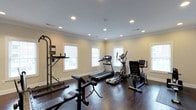 This fitness center is a thoughtfully designed single-floor workout space that combines functionality with a clean, inviting aesthetic. The room features rich, dark hardwood flooring that provides a durable and attractive surface suited for a variety of exercises. Crown molding lines the perimeter of the smooth, neutral-toned ceiling, which is outfitted with recessed lighting to ensure consistent, well-distributed illumination throughout the space.

Multiple windows along the walls allow natural light to flow in, creating a bright and energizing atmosphere. The windows are fitted with blinds for adjustable light control and privacy.

The fitness center is equipped with a well-rounded selection of exercise equipment, including a treadmill, an elliptical trainer, and a stationary recumbent bike for cardiovascular workouts. A multi-function strength training tower provides options for pull-ups, dips, and other bodyweight exercises, while a dedicated weight bench supports resistance and strength training routines.

The layout is open and spacious, allowing comfortable movement between equipment. The neutral wall tones and clean architectural detailing contribute to a focused and motivating environment, making this fitness center a practical and well-appointed space for a comprehensive workout experience.