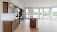 This single-floor unit presents a clean, contemporary design characterized by a neutral palette of whites and warm wood tones. The open-concept kitchen and living area serves as the heart of the space, where rich dark-wood cabinetry lines the kitchen walls alongside stainless steel appliances, including a range, microwave, and refrigerator. A central island with a white countertop and pendant lighting above provides additional workspace and casual seating. Large floor-to-ceiling windows flood the area with natural light and offer views of the surrounding neighborhood, enhancing the sense of openness. Light hardwood flooring extends throughout the main living areas, contributing to a warm, cohesive aesthetic.

The bedrooms feature plush carpeting underfoot and generous windows that continue the theme of abundant natural light. One carpeted bedroom includes a wall-mounted thermostat and baseboard ventilation, while another boasts a large picture window with exterior views.

Two bathrooms serve the unit, each finished with light hardwood-style flooring and dark wood vanity cabinetry paired with white countertops. One bathroom includes a tub-shower combination, while the other offers a walk-in shower configuration with a tall storage cabinet. Both feature well-lit vanity mirrors. Overall, the unit presents a polished, move-in-ready environment with consistent modern finishes throughout.