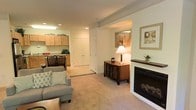 This single-floor residence offers a thoughtfully designed layout that balances comfort and functionality. The living area is a welcoming focal point, anchored by a classic white-framed fireplace with a dark wood mantel, providing both visual warmth and a cozy atmosphere. Soft carpeting, recessed lighting, and neutral wall tones contribute to the space's calm and inviting ambiance. A stylish sideboard with decorative accents and framed wall art add a refined finishing touch to the living area.

The open flow of the layout connects seamlessly to the kitchen and dining area. The kitchen features light wood cabinetry with ample storage, recessed and overhead lighting, and a clean, efficient workspace. A dining set positioned nearby creates a natural and convenient space for everyday meals or entertaining.

A set of double doors along the interior hallway suggests additional storage or closet space, enhancing the home's practical appeal. Structural beam detailing on the ceiling adds a subtle architectural character throughout the main living zone.

Overall, the space presents a harmonious blend of traditional design elements and everyday livability, making it a comfortable and well-organized home.