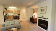 This single-floor residence offers a thoughtfully designed layout that balances comfort and functionality. The living area is a welcoming focal point, anchored by a classic white-framed fireplace with a dark wood mantel, providing both visual warmth and a cozy atmosphere. Soft carpeting, recessed lighting, and neutral wall tones contribute to the space's calm and inviting ambiance. A stylish sideboard with decorative accents and framed wall art add a refined finishing touch to the living area.

The open flow of the layout connects seamlessly to the kitchen and dining area. The kitchen features light wood cabinetry with ample storage, recessed and overhead lighting, and a clean, efficient workspace. A dining set positioned nearby creates a natural and convenient space for everyday meals or entertaining.

A set of double doors along the interior hallway suggests additional storage or closet space, enhancing the home's practical appeal. Structural beam detailing on the ceiling adds a subtle architectural character throughout the main living zone.

Overall, the space presents a harmonious blend of traditional design elements and everyday livability, making it a comfortable and well-organized home.