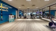 Located at 2101 Market St in Denver, Colorado, this contemporary fitness center offers a thoughtfully designed, single-floor space built to support a wide range of workout routines. The facility is finished with light wood-look flooring throughout, crisp white ceilings with recessed lighting, and striking bold blue accent walls that give the space an energetic, modern character.

A dedicated spin studio anchors one side of the facility, lined with a row of stationary cycling bikes equipped with individual digital screens. Large framed mirrors span the studio walls, amplifying the sense of space and allowing members to monitor their form. The studio is accessible through a glass-paneled door branded with the building's "21" logo, maintaining visual connectivity with the broader fitness floor.

The main workout area is generously open, providing ample room for functional training and group exercise. Equipment is thoughtfully organized along the perimeter, including a standing punching bag, medicine balls, stability and exercise balls on a dedicated rack, kettlebells, and yoga mats stored neatly in a built-in cubby shelf. The glass partition walls between zones allow natural sightlines across the entire facility, creating a cohesive and inviting atmosphere. Overall, the space strikes a balance between professional amenity and approachable design.