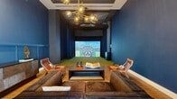 Located at 700 Northeast 1st Avenue in the heart of Miami, this purpose-built entertainment room delivers an upscale, immersive recreational experience within a single, thoughtfully designed space. The room is defined by its deep navy blue walls accented with classic wainscoting paneling, creating a sophisticated and intimate atmosphere. Warm wood-look flooring runs throughout the lounge area, offering a striking contrast to the bold wall color.

At the heart of the space is a state-of-the-art simulator bay, complete with artificial turf matting and a large projection screen capable of displaying golf courses and dart games, making it a versatile hub for entertainment. Dark floor-to-ceiling curtains flank the simulator bay, enhancing the immersive experience by minimizing ambient light during play.

The lounge seating area is furnished with a plush navy sofa, cognac leather accent chairs, and a low-profile rectangular coffee table, providing comfortable viewing and social space directly facing the simulator. A sculptural brass and amber glass sputnik chandelier hangs overhead, casting warm ambient light that complements the room's refined aesthetic. A decorative credenza with an artistic sculpture and a tall column floor lamp complete the interior, adding curated detail to the overall design.