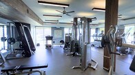 This fitness center offers a thoughtfully designed workout environment suited for a full range of exercise routines. The single-floor layout makes efficient use of space, housing an impressive selection of equipment including a multi-station cable machine, weight benches, free weights, and cardio machines such as treadmills visible along the mirrored wall. The mirrored walls serve a dual purpose — aiding exercisers in monitoring their form while also visually expanding the room and enhancing its open feel. Ceiling fans are mounted overhead, contributing to airflow and comfort during workouts, while hanging fluorescent lighting ensures the space is well-lit throughout. Large windows line one side of the room, flooding the interior with natural light and offering views of the surrounding outdoor greenery, creating an inviting and energizing atmosphere. The flooring features a durable rubber surface, ideal for high-traffic fitness use and providing cushioning underfoot. The walls are painted in a calm, neutral blue-gray tone that lends the space a clean, focused ambiance. Decorative wooden beam accents add a subtle touch of warmth to the otherwise modern, functional aesthetic, making this fitness center both practical and visually appealing.