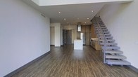 This contemporary two-floor space showcases a thoughtfully designed open-concept layout that seamlessly connects the living and kitchen areas. Rich, dark hardwood-style flooring runs throughout the main level, grounding the space with warmth and contrast against the clean, light grey walls and white ceilings. Recessed lighting is embedded throughout the ceiling, providing even, modern illumination across the open floor plan.

The kitchen is a standout feature, outfitted with warm wood cabinetry, stainless steel appliances, and a sleek white kitchen island that serves as both a functional workspace and a visual centerpiece. A statement range hood anchors the cooking area, adding an industrial-modern touch that complements the overall aesthetic.

A striking open-riser staircase with a metal frame and wood treads rises along the right side of the main floor, offering a sculptural architectural element while providing access to the second level. The entryway, framed by a bold dark grey door, adds a strong focal point at the rear of the main floor.

The space benefits from a generous ceiling height on the main level, contributing to an airy, expansive atmosphere. Overall, the design strikes a balance between modern industrial elements and warm, residential finishes, creating a refined and versatile environment.