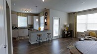 This single-floor space is designed with an open-concept layout that seamlessly connects the kitchen and living areas, creating a cohesive and inviting environment. The kitchen is outfitted with crisp white cabinetry, granite countertops, and a peninsula with bar seating for two, making it both functional and ideal for casual dining or entertaining. A track lighting fixture illuminates the kitchen workspace, while stylish pendant lights hang above the peninsula, adding a contemporary decorative touch. The kitchen also features a full suite of appliances and a sink positioned beneath a window, allowing for natural light during food preparation. Neutral gray walls throughout complement the rich, dark hardwood-style flooring that extends across the entire space, lending warmth and visual continuity. Multiple windows fitted with blinds and patterned valances allow generous amounts of natural light to fill the interior, enhancing the open and airy feel. A glass-panel door provides additional light and access to the exterior. The living area flows naturally from the kitchen, offering a versatile and well-lit space. Decorative accents, including wall-mounted circular pieces and warm-toned accessories, add character throughout. Overall, the space offers a clean, modern aesthetic with thoughtful design details.