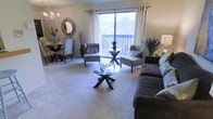 This single-floor residence seamlessly blends the living and dining areas into one cohesive, open layout. The living area is anchored by a plush, dark upholstered sofa paired with textured accent chairs and a distinctive geometric glass coffee table, creating a comfortable and stylish seating arrangement. Neutral-toned carpeting runs throughout, contributing to the warm, welcoming ambiance of the space.

A large sliding glass door dominates one wall, flooding the interior with natural light and providing direct access to a private balcony that overlooks lush, tree-lined surroundings. White curtain panels frame the door, softening the transition between indoors and out.

The dining area is positioned adjacent to the living space and features an elegant round table with ornate chairs, complemented by a decorative sunburst mirror and a chandelier overhead, adding a refined touch to the overall aesthetic. Carefully placed greenery and decorative accents throughout reinforce the polished, curated feel of the space. Together, the thoughtful furnishings, natural light, and outdoor views create a balanced and comfortable living environment.