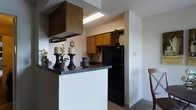This single-floor space offers a practical and inviting layout centered around a well-appointed kitchen. The kitchen is a standout feature, showcasing warm wood cabinetry that provides ample storage alongside sleek dark countertops that add a touch of contrast and sophistication. A peninsula-style breakfast bar serves as a natural divider between the kitchen and the adjacent living area, making it ideal for casual dining or entertaining. The black refrigerator and overhead range hood contribute to a cohesive, functional aesthetic. A textured ceiling adds a classic touch throughout the space, while neutral wall tones keep the atmosphere light and versatile. Decorative accents, including candle holders and potted plants, hint at the warmth and character that can be brought to the space. The kitchen transitions smoothly into the surrounding areas, with glimpses of a carpeted living zone and a dining area just at the periphery. The overall ambiance is cozy and efficient, making thoughtful use of the available footprint. This space is well-suited for those seeking a comfortable, easy-to-maintain environment with a functional kitchen at its heart.