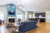 Located at 32 Ashbury Woods Dr in Huntsville, Alabama, this single-story community clubhouse offers a well-appointed, open-concept layout designed to serve both social and professional needs. The centerpiece of the space is a spacious great room anchored by soaring vaulted ceilings and an elegant chandelier, drawing the eye toward a teal accent wall that frames a wall-mounted television above a classic white fireplace. Arched windows flank the focal wall, flooding the room with natural light, while warm hardwood flooring runs throughout the entire space, unifying each area seamlessly. Decorative white columns with paneled bases define the transitions between zones without interrupting the open flow. A dedicated leasing or office area is thoughtfully positioned near the entrance, furnished with a full-size desk and guest seating. A dining nook features a charming arched window overlooking the outdoors, providing an intimate setting for small meetings or consultations. The full kitchen is outfitted with rich espresso cabinetry, dark granite countertops, a tile backsplash, and ample counter space accommodating a printer and office equipment, reflecting a dual-purpose utility. Multiple glass entry doors at the rear invite natural light and provide easy outdoor access, completing this functional and welcoming community facility.