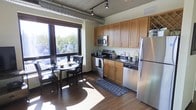 This single-floor, two-bedroom, two-bathroom unit showcases a distinctive industrial-loft design, characterized by exposed silver ductwork along the ceilings, dark-trimmed oversized windows, and rich hardwood flooring throughout. The open layout maximizes the sense of space, allowing natural light to flow generously across the interior.

The living area is anchored by large picture windows that frame verdant treetop views, creating a bright and airy atmosphere. The space comfortably accommodates a seating arrangement and media center, with track lighting overhead providing additional illumination.

The kitchen is well-appointed with warm honey-toned cabinetry extending to the ceiling for ample storage, stainless steel appliances including a refrigerator, stove, and built-in microwave, and a dark stone countertop. A dedicated dining area positioned beside a generous window enjoys abundant natural light, making it an inviting spot for meals.

Throughout the unit, the consistent use of dark hardwood flooring, industrial ceiling details, and large windows creates a cohesive, modern urban aesthetic that balances both functionality and style.