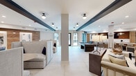 This expansive amenity lounge is thoughtfully designed to accommodate a variety of social and recreational activities within a single, open-plan floor. The space is anchored by a striking oversized kitchen island topped with elegant white marble-veined countertops, suspended above by a trio of gold disc pendant lights that add a warm, contemporary flair. The kitchen area features dark cabinetry, a stainless steel sink, and a wood-accented feature wall with a mounted flat-screen TV, creating an ideal setting for gatherings. Adjacent to the kitchen, a billiards table and shuffleboard court offer dedicated spaces for recreation and entertainment. The lounge is punctuated by structural columns and dark exposed ceiling beams that add architectural character against the crisp white ceiling. Large windows allow natural light to fill the space, offering views to an outdoor courtyard. Throughout the room, distinct seating zones are defined by high-backed upholstered booth-style sofas, plush lounge chairs, and a round dining table with velvet accent chairs, all set atop large-format light tile flooring. Warm wood wall cladding, textured brick accent walls, and curated artwork contribute to the inviting, upscale ambiance of the space.