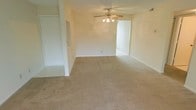 This single-floor space presents a clean, functional layout centered around a generously sized main living area. The room is finished with wall-to-wall beige carpeting that adds warmth underfoot and complements the neutral, cream-toned walls throughout. A ceiling fan with an integrated light fixture is centrally mounted, providing both air circulation and ambient lighting to the space. The open design of the living area allows for flexible furniture arrangement, making it adaptable to various needs. A doorway on the left side of the room leads to what appears to be a closet or storage area, while an open passage on the right provides access to an adjacent room, contributing to the overall flow of the floor plan. The layout feels connected and cohesive, with consistent finishes carried throughout. White baseboards frame the carpeted floors, adding a clean, finished look. The overall ambiance is simple and neutral, offering a blank canvas that is well-suited for personalizing with furnishings and décor.