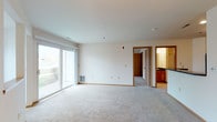This single-floor unit is designed with a practical, open layout that connects the living and kitchen areas seamlessly. The spacious living room features neutral-toned carpet, warm wood baseboards, and a large sliding glass door that fills the space with natural light while providing direct access to an outdoor patio. A peninsula-style breakfast bar separates the kitchen from the living area, where warm oak cabinetry, dark granite-style countertops, and black appliances—including a range, built-in microwave, and refrigerator—create a functional and cohesive cooking space. Light hardwood flooring runs throughout the kitchen, complementing the wood-toned cabinetry.

The unit includes multiple bedrooms, each carpeted and finished with neutral walls, wood baseboards, and windows offering views of surrounding greenery. One bedroom features a built-in shelving niche, adding a distinctive architectural detail. The bathroom is well-appointed with oak vanity cabinetry, dark countertops, a large mirror with overhead lighting, and a full soaking tub with a shower. Ceiling-mounted light fixtures illuminate the common areas, and the overall ambiance is clean, bright, and move-in ready.