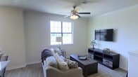 This single-story space showcases a well-appointed living area designed for comfort and practicality. The room features wood-style flooring that adds warmth and durability underfoot, complemented by light-toned walls that create a bright, open atmosphere. A large multi-pane window allows generous natural light to fill the space, providing views of the surrounding greenery outside.

The living area is anchored by a ceiling fan with an integrated light fixture, ensuring year-round comfort and adequate illumination. Crown molding runs along the upper edges of the walls, adding a refined architectural detail to the room. A visible air vent near the ceiling further highlights the functional infrastructure integrated throughout the space.

The layout comfortably accommodates a full seating arrangement, including a sofa paired with accent pillows and a throw, centered around a dark-toned wooden coffee table. Opposite the seating, a media console in a matching dark finish provides ample storage and display space, ideal for entertainment setups. The overall ambiance is casual yet polished, balancing everyday functionality with thoughtful design details.
