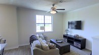 This single-story space showcases a well-appointed living area designed for comfort and practicality. The room features wood-style flooring that adds warmth and durability underfoot, complemented by light-toned walls that create a bright, open atmosphere. A large multi-pane window allows generous natural light to fill the space, providing views of the surrounding greenery outside.

The living area is anchored by a ceiling fan with an integrated light fixture, ensuring year-round comfort and adequate illumination. Crown molding runs along the upper edges of the walls, adding a refined architectural detail to the room. A visible air vent near the ceiling further highlights the functional infrastructure integrated throughout the space.

The layout comfortably accommodates a full seating arrangement, including a sofa paired with accent pillows and a throw, centered around a dark-toned wooden coffee table. Opposite the seating, a media console in a matching dark finish provides ample storage and display space, ideal for entertainment setups. The overall ambiance is casual yet polished, balancing everyday functionality with thoughtful design details.