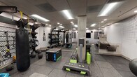 Located at 310 E Sixth St in Tucson, AZ, this single-floor fitness center is designed to accommodate a wide range of workout styles and training needs. The space features a thoughtfully designed interior with a distinctive aesthetic, highlighted by perforated white wall panels accented with a graduating dot pattern that adds visual depth and a modern, industrial character throughout.

The main training floor is surfaced with durable rubber flooring in a dark tile format, providing both safety and performance underfoot. The area is well-equipped with functional training tools including plyo boxes, step platforms, foam rollers, battle ropes, TRX suspension trainers, kettlebells, medicine balls, and a heavy punching bag mounted to a freestanding multi-functional rack. A pegboard wall organizes additional accessories and free weights for efficient access.

Large wall-mounted mirrors expand the perceived scale of the space and allow users to monitor their form during workouts. Recessed and panel lighting throughout the drop ceiling ensures bright, even illumination across the entire floor. A large flat-screen Samsung display and a mounted speaker system are integrated into the wall, supporting class instruction or entertainment.

A water fountain station is visible near the mirrored section, and a dedicated entry door with a small vestibule provides a clean transition into the space.